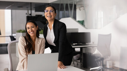 Young and mature professional, multiethnic women posing in workplace, look at camera with smile, enjoy workflow, feeling motivation, satisfied with productive teamwork in coworking office. Mentorship