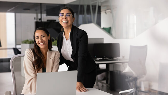Young and mature professional, multiethnic women posing in workplace, look at camera with smile, enjoy workflow, feeling motivation, satisfied with productive teamwork in coworking office. Mentorship - Powered by Adobe