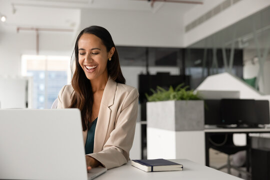 Happy woman typing on laptop keyboard sits at desk in coworking office, feel satisfied while exchange e-mails with client or colleague, assisting remotely, working online on project or presentation