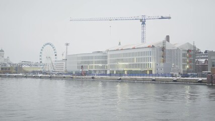 A waterfront scene showcasing a construction site amidst winter, featuring a crane, new buildings, and a Ferris wheel in the background, highlighting urban development and seasonal beauty.