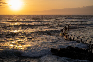 The ocean is calm and the sun is setting. A man is standing on a pier. The water is choppy and the sky is orange