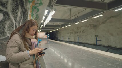 A young individual waits at a subway station, absorbed in their phone, surrounded by urban artwork. This captures a blend of modern lifestyle and daily commute.