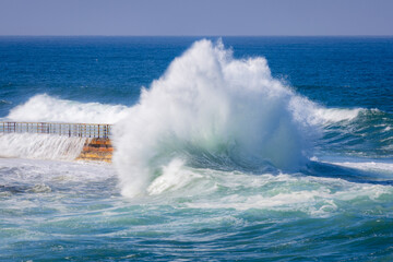 Big powerful king tide waves crashing and exploding onto a wall in San Diego Southern California during the day with a teal blue ocean in the background