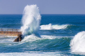 Fototapeta premium Big powerful king tide waves crashing and exploding onto a wall in San Diego Southern California during the day with a teal blue ocean in the background