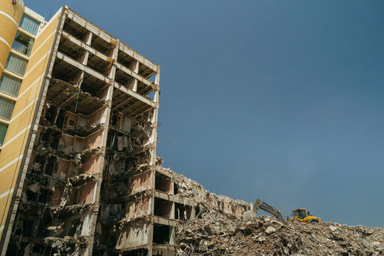 Collapsed building with an excavator clearing heavy rubble at a dusty demolition site in an urban danger zone. Partially destroyed high-rise building after earthquake or explosion. Copy space