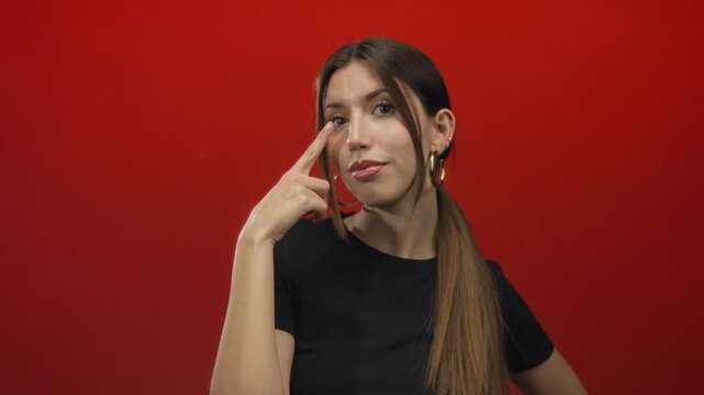 Woman points finger to eye in studio with red backdrop wearing hoop earrings and black top; focus clarity.