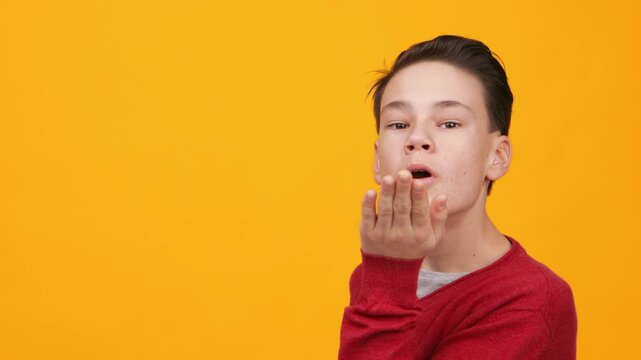 A young boy with short hair is blowing a kiss towards the camera. He wears a red sweater and is set against a bright yellow background. The scene captures a joyful moment.