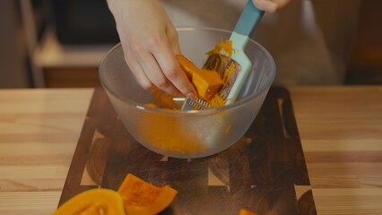 Focus on female hands grating fresh butternut squash into a glass bowl. Blurred kitchen background and squash slices on wooden cutting board in the foreground. Home cooking process, healthy lifestyle.