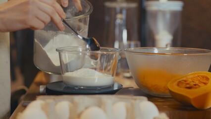 Woman adding flour to pancake batter in a glass bowl on scales. Grated butternut squash and a half pumpkin on a wooden table. Process of making healthy pumpkin pancakes in a cozy home kitchen setting.