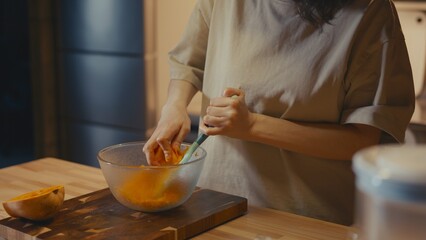 Medium shot of woman grating fresh butternut squash in a home kitchen. Half of the squash lies on wooden cutting board. Blurred domestic background, warm atmosphere, healthy organic meal preparation.