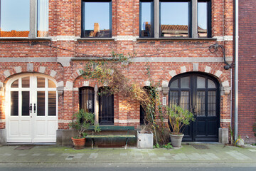 Historic European brick townhouse facade in Bruges with arched doors, black windows and leafy plants on sidewalk in afternoon sunlight