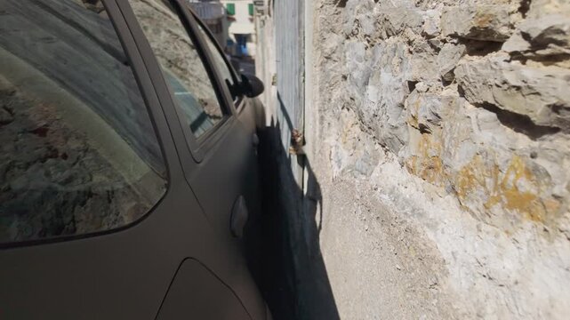 Black car parked extremely close to an old wall. Experienced driver shows precise parking on a narrow Mediterranean street. Dolly out shot reveals the tiny space between vehicle and wall.