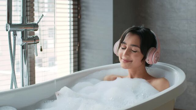 A woman enjoys a bath full of bubbles while wearing headphones in a modern bathroom with large windows letting in sunlight. She appears happy and relaxed during her personal time.