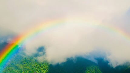 Naklejka premium A colorful rainbow spans across the sky above verdant mountains, partially obscured by clouds, showcasing a beautiful natural landscape with vivid hues