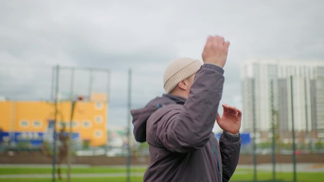 man shadowboxing in urban park, wearing beanie and jacket, overcast sky and residential skyline in background, performing punching combinations and warmup drills, solo fitness routine, determined