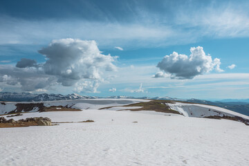 Scenic landscape with sunlit snowfield against snow cornice and big snowbound mountain range far away under dramatic cloudy sky. Vast view with snowy glade in high mountains under clouds in blue sky.