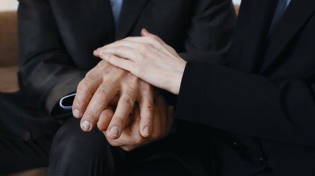 Two individuals in suits are seated closely together, holding hands. The scene shows a moment of connection in a bright indoor environment. Their expressions show engagement in discussion.