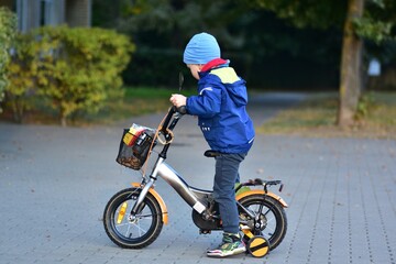 Toddler standing beside a small bicycle with training wheels, adjusting position before riding on a paved path. Childhood independence, learning balance and outdoor activity in a calm park setting.