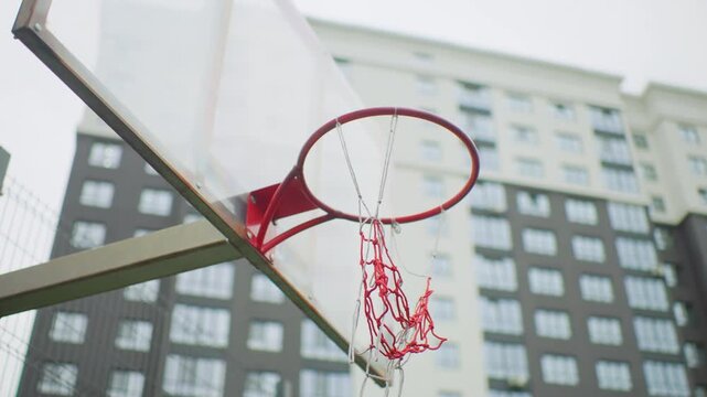 urban basketball hoop close up red rim, transparent backboard with chain net, highrise apartment backdrop, overcast sky, lowangle perspective, motion implied ball trajectory, weathered paint