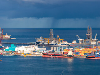 Fototapeta premium Container stacks, tank farms, and large vessels occupy the Port of Las Palmas, Gran Canaria, Spain. Two offshore drilling platforms stand tall as storm clouds pass.