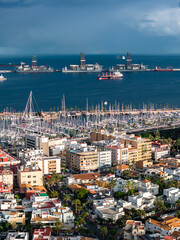 Fototapeta premium Aerial view shows Las Palmas de Gran Canaria marina and port, yachts and masts packed by the waterfront, drilling platforms and cargo ships beyond under dramatic light.