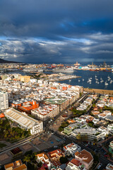 Fototapeta premium Aerial view shows Las Palmas de Gran Canaria harbor, Puerto de La Luz cranes, marina sailboats, stadium like structure, gridded streets, and sunlit homes under storm clouds.