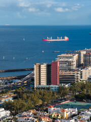 Fototapeta premium Las Palmas de Gran Canaria port scene shows mid and high rises, a red cargo ship offshore, curving breakwaters, sailboats, tennis courts, and palm lined streets in soft afternoon light.