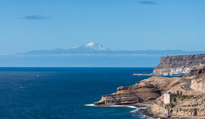 Southwest Gran Canaria shows rugged cliffs, the GC 500 road, a small harbor near Puerto de Mogan, and Mount Teide on Tenerife on the horizon in bright daytime light. © Aerial Film Studio