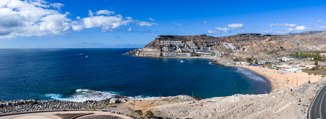 Aerial panorama shows Puerto Rico de Gran Canaria and Amadores Bay, golden sand beneath terraced cliffs, turquoise water, sailboat, road, breakwater, midday light.