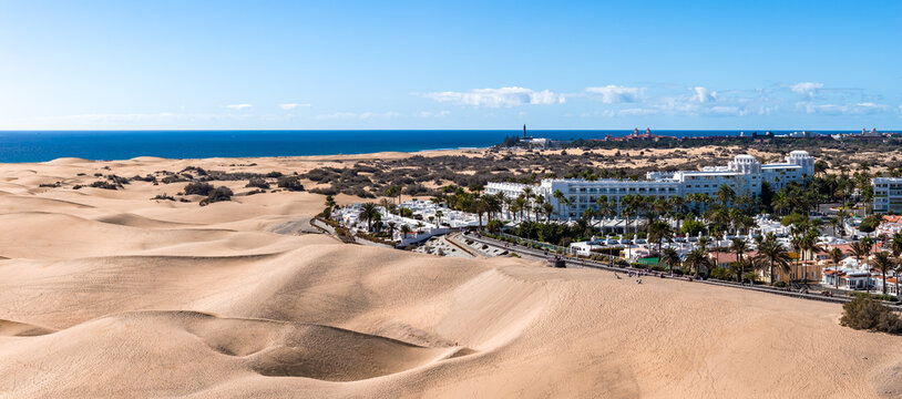 Maspalomas Dunes in Gran Canaria stretch to the Atlantic. Rippling sand hills, Maspalomas Lighthouse on the horizon, white resort buildings, and palm lined streets in bright midday light.
