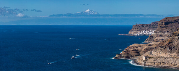 Terraced Teide volcanic cliffs and a curving road meet a bay town on Gran Canaria. Boats leave white wakes on deep water, Mount Teide rises beyond in clear winter light. © Aerial Film Studio