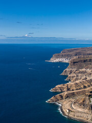 Aerial daylight view of Gran Canaria southwest coast, volcanic cliffs, coastal road, whitewashed resorts, and boats. Mount Teide on Tenerife rises above a cloud band. © Aerial Film Studio
