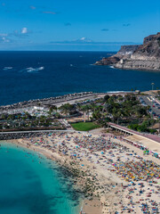 Fototapeta premium Aerial scene shows Amadores Beach, Gran Canaria, with umbrellas, promenade, palm park, marina, rocky headlands, and distant Mount Teide under bright midday light.