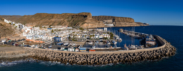 Aerial view of Puerto de Mogan on Gran Canaria shows a calm marina, sailboats, concrete tetrapods on the breakwater, whitewashed buildings, palms, and rugged ochre cliffs. © Aerial Film Studio