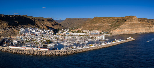 Aerial view shows Puerto de Mogan, Gran Canaria, marina with masts and yachts, concrete tetrapods breakwater, whitewashed canals, palm streets, ochre cliffs, small boat departing. © Aerial Film Studio
