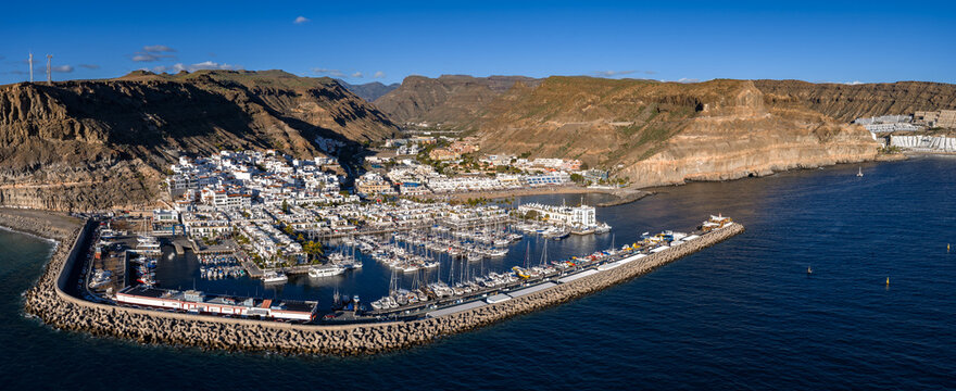 Aerial view of Puerto de Mogan, Gran Canaria, with a semicircular breakwater, packed marina, whitewashed buildings, canals, and ochre cliffs under clear midday skies.