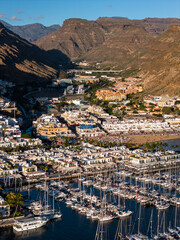 Aerial view of Puerto de Mogan on Gran Canaria shows a packed marina with sailboats, whitewashed buildings, a sandy beach with palms, a viaduct, and terraced hillsides. © Aerial Film Studio