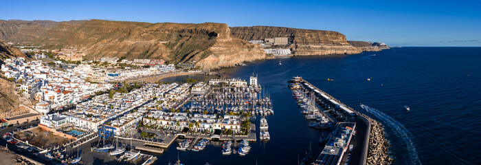 Aerial view shows Puerto de Mogan on Gran Canaria, with whitewashed houses, canals, a yacht marina, sandy beach, and breakwater under bright midday light and clear sky. © Aerial Film Studio