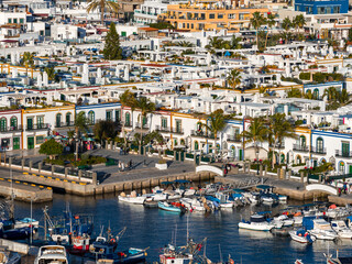 Whitewashed buildings with colorful frames, arched bridges, and canals define Puerto de Mogan in Gran Canaria, where boats line a calm harbor and palms frame a sunlit promenade. © Aerial Film Studio