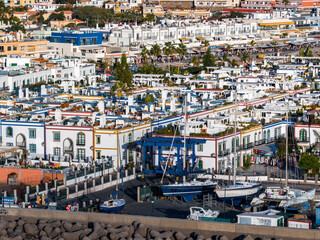 Puerto de Mogan on Gran Canaria shows a marina quarter with a gantry crane lifting yachts, whitewashed buildings, palm lined promenades, and people dining and strolling. © Aerial Film Studio