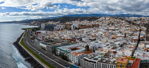 Aerial view shows Las Palmas de Gran Canaria seafront, coastal highway, whitewashed hills, Cathedral of Santa Ana, mid rise blocks, mountains, clouds, traffic, and surf. © Aerial Film Studio