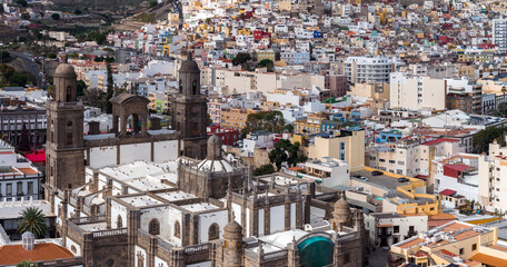 Catedral de Santa Ana dominates the old quarter in Las Palmas de Gran Canaria, twin bell towers and dark stone facade stand above tight rooftops in soft daylight. © Aerial Film Studio