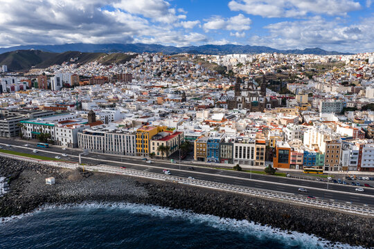 Aerial coastal skyline of Las Palmas de Gran Canaria shows rocky shore, seafront road, low rise buildings, and twin towered Cathedral of Santa Ana under soft light and clouds.