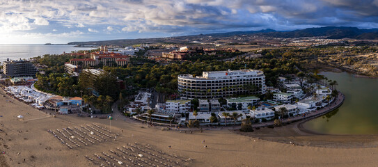 Fototapeta premium A wide aerial view shows Maspalomas, Gran Canaria, with dunes, La Charca curving right, the Palm Beach hotel centered, terracotta hotels, palm groves, and late afternoon clouds.