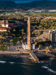 Fototapeta premium Aerial Maspalomas Lighthouse rises by a seafront promenade in Gran Canaria, Spain. Late afternoon light casts long shadows over parks, resorts, a breakwater, and distant mountains.