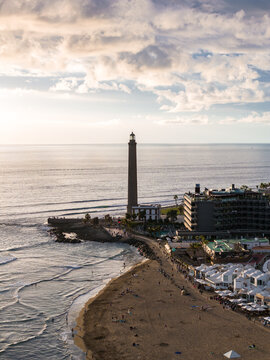 Aerial view shows Maspalomas Lighthouse on a rocky point beside a sandy beach in Gran Canaria, Canary Islands. Late afternoon light casts long shadows over calm surf.