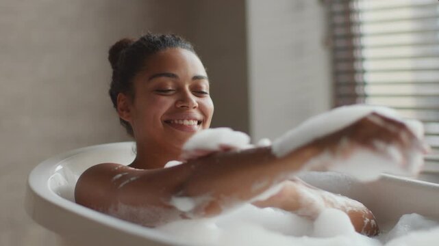A woman sits in a bathtub filled with bubbles, smiling as she plays with the foam. Soft light filters through window blinds, creating a cozy atmosphere in the room.