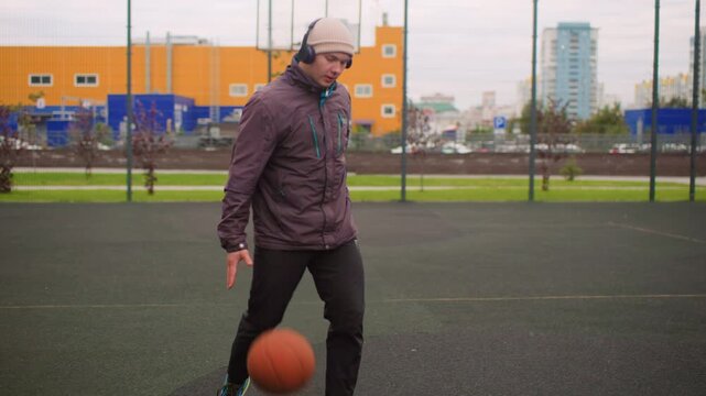 solo man dribbling basketball outdoors on urban court wearing beanie and headphones, layered jacket and sneakers, focused training routine under overcast sky with city buildings in background,