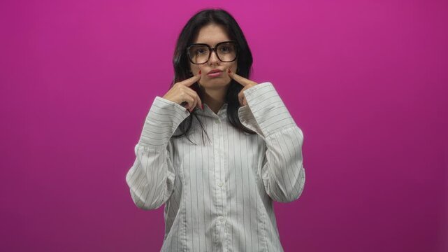 Young hispanic woman pushes cheeks outward with both index fingers in bright pink studio setting; toothache discomfort.