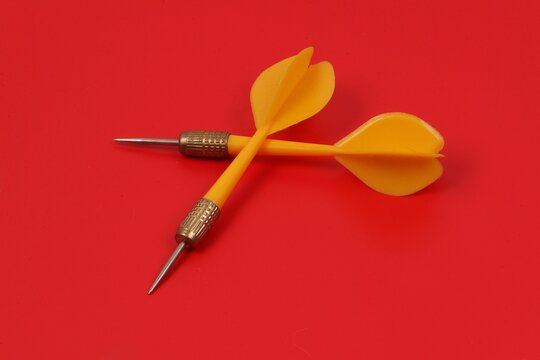 Close-up of two darts with a metal tip and yellow plastic grip on a red background